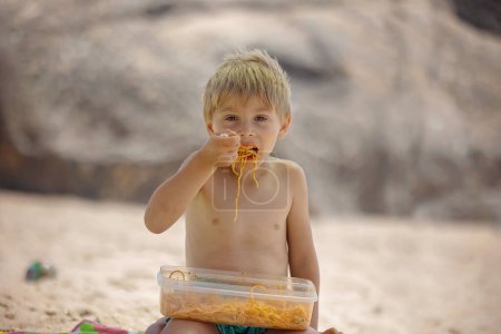 Foto de Hermoso niño rubio, chico, comiendo espaguetis en la playa de Portugal en un día nublado de niebla, verano - Imagen libre de derechos