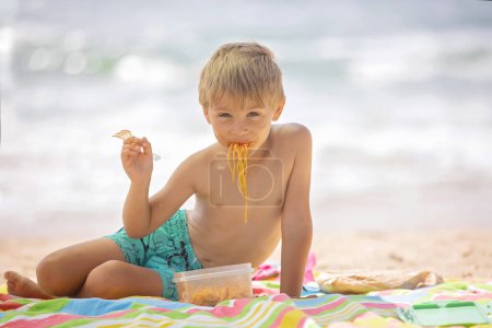 Foto de Hermoso niño rubio, chico, comiendo espaguetis en la playa de Portugal en un día nublado de niebla, verano - Imagen libre de derechos