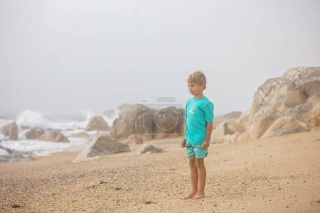Foto de Hermoso niño rubio, muchacho, la recolección de conchas en la playa de Portugal en un día nublado niebla, verano - Imagen libre de derechos