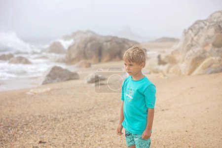 Téléchargez les photos : Belle enfant blonde, garçon, ramassant des coquillages sur la plage au Portugal par une journée nuageuse brumeuse, l'été - en image libre de droit