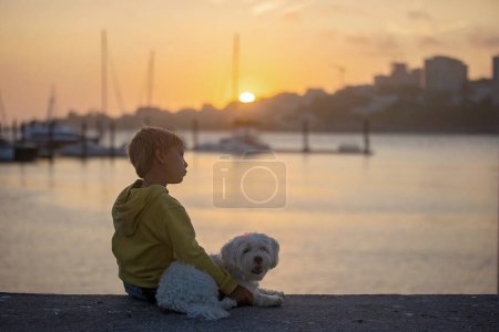 Foto de Niños, niños, hermanos, disfrutando del atardecer sobre el río con su perro y mamá malteses, barcos, sol, río, océano - Imagen libre de derechos