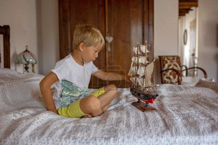 Téléchargez les photos : Enfant blond mignon, garçon, jouant à la maison avec des jouets, bateau, poupées, maison autentique - en image libre de droit
