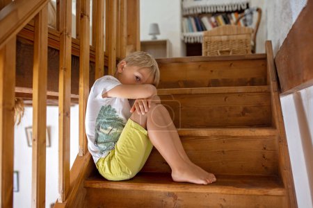 Téléchargez les photos : Enfant blond mignon, garçon, assis sur un escalier, triste et solitaire, maison autentique - en image libre de droit