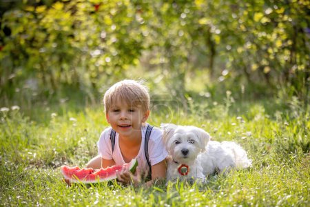 Téléchargez les photos : Incroyable enfant blond en bas âge, garçon avec chien, manger de la pastèque dans le jardin, l'été - en image libre de droit