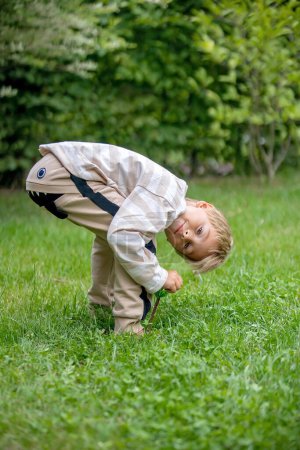 Téléchargez les photos : Petit enfant d'âge préscolaire, avec des chiffons drôles, faisant sourire le visage, regardant à l'envers entre ses jambes dans le jardin - en image libre de droit