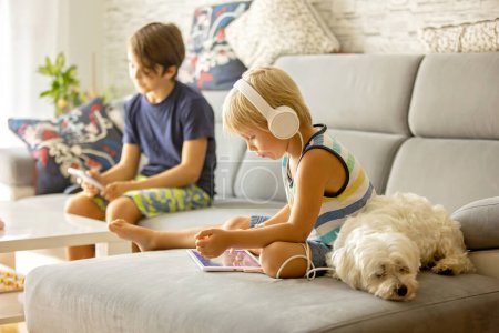 Téléchargez les photos : Mignon enfant, garçon, regarder le film sur tablette à la maison, chien maltais mignon couché à côté de lui sur le canapé - en image libre de droit