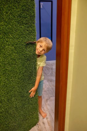 Téléchargez les photos : Mignon blond tout-petit enfant, higing derrière la porte à la maison, regardant curieusement et souriant - en image libre de droit