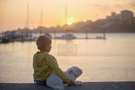 Téléchargez les photos : Enfants, garçons, frères, profiter du coucher de soleil sur la rivière avec leur chien maltais animal de compagnie et maman, bateaux, soleil, rivière, océan - en image libre de droit