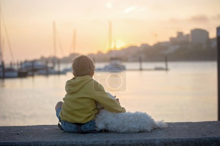 Foto de Niños, niños, hermanos, disfrutando del atardecer sobre el río con su perro y mamá malteses, barcos, sol, río, océano - Imagen libre de derechos