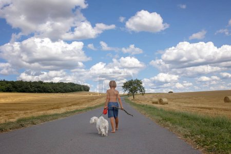 Foto de Hermoso niño rubio, muchacho, caminando por el camino rural con su dulce perrito maltés. Increíble paisaje, escena rural con nubes, árbol y camino vacío verano, campos de pajar al lado de la carretera, Portugal - Imagen libre de derechos