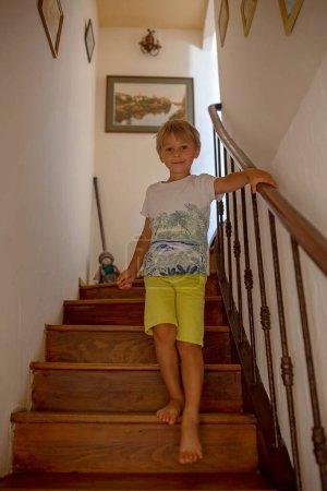Foto de Pequeño niño triste, niño, sentado triste en una escalera en casa, castigado, vieja casa autentica - Imagen libre de derechos