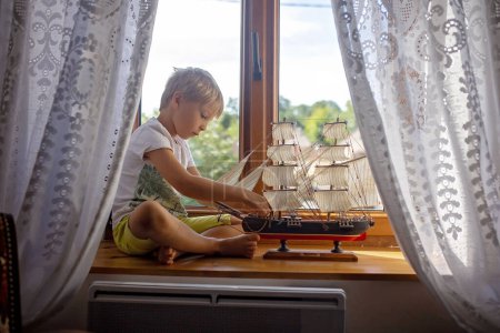 Téléchargez les photos : Mignon enfant blond, garçon, jouer à la maison avec un gros navire sur la fenêtre de la vieille maison autentique - en image libre de droit