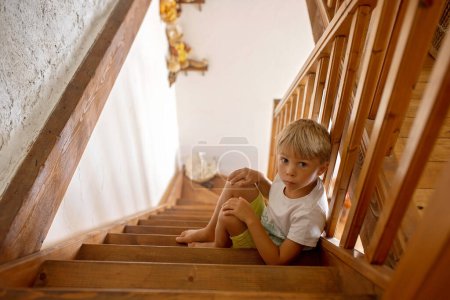 Téléchargez les photos : Enfant blond mignon, garçon, assis sur un escalier, triste et solitaire, maison autentique - en image libre de droit
