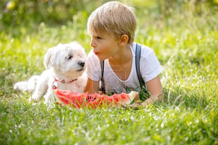 Foto de Increíble niño rubio, niño con perro de compañía, comer sandía en el jardín, verano - Imagen libre de derechos