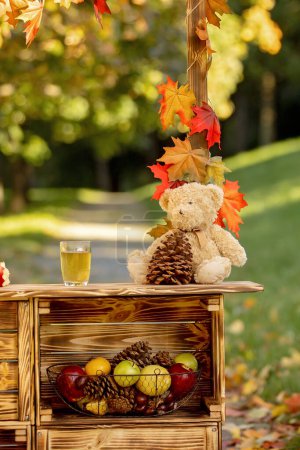 Foto de Niño rubio lindo, de pie junto al soporte de madera de otoño con decoración, manzanas, hojas, taza, erizo en el parque, otoño - Imagen libre de derechos