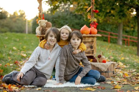 Foto de Lindo niño rubio y hermanos hermanos, de pie junto al soporte de madera de otoño con decoración, manzanas, hojas, taza, erizo en el parque, otoño - Imagen libre de derechos