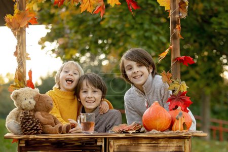 Foto de Lindo niño rubio y hermanos hermanos, de pie junto al soporte de madera de otoño con decoración, manzanas, hojas, taza, erizo en el parque, otoño - Imagen libre de derechos