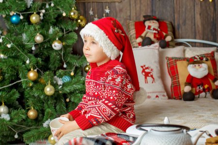 Téléchargez les photos : Enfant tout-petit mignon, garçon en tenue de Noël, jouant dans une cabane en bois à Noël, décoration autour de lui. Livre pour enfants et thé à boire - en image libre de droit