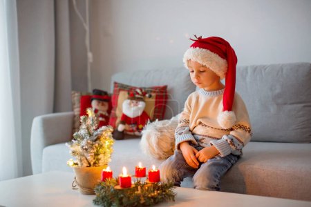Foto de Lindo niño pequeño, niño con un traje de Navidad, jugando en una cabaña de madera en Navidad, decoración a su alrededor. Libro de lectura infantil y beber té - Imagen libre de derechos