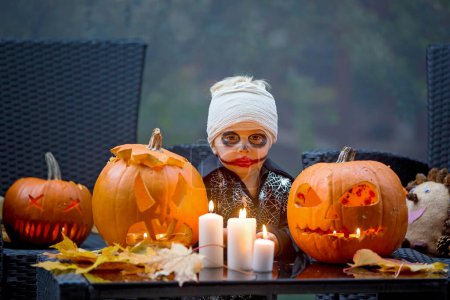 Téléchargez les photos : Enfant effrayant en costume d'Halloween, jouant avec des citrouilles et des bougies sculptées - en image libre de droit