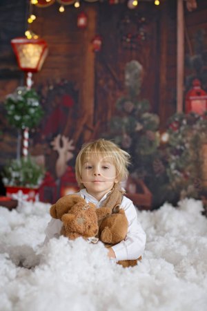 Foto de Lindo niño de moda, jugando en la nieve con oso de peluche en frente de un registro de cabaña de madera, decoración de Navidad - Imagen libre de derechos