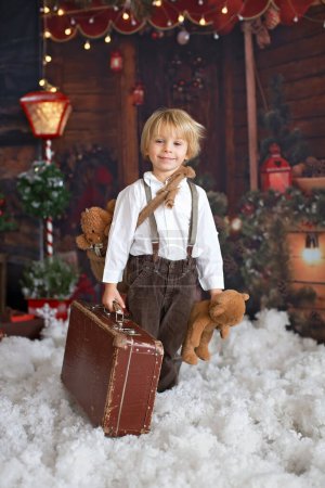 Foto de Lindo niño de moda, jugando en la nieve con oso de peluche en frente de un registro de cabaña de madera, decoración de Navidad - Imagen libre de derechos