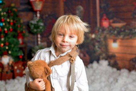 Téléchargez les photos : Bébé mignon garçon de mode, jouer dans la neige avec ours en peluche devant une bûche de cabine en bois, décoration de Noël - en image libre de droit