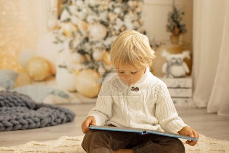 Foto de Lindo niño rubio preescolar, leyendo un libro y regalo de apertura en Navidad en casa acogedora, luces y decoración - Imagen libre de derechos