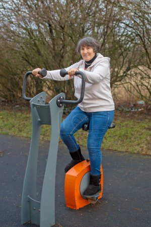 Téléchargez les photos : Femme d'âge moyen, faisant de l'exercice dans le parc, faisant du vélo dans un centre de remise en forme extérieur, hivernant - en image libre de droit