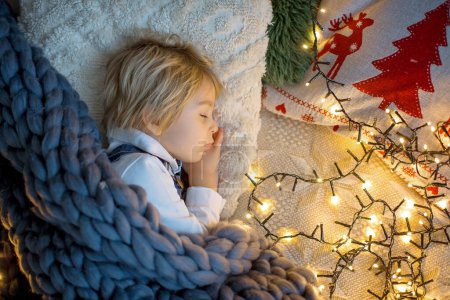 Photo for Toddler child, cute blond boy, sleeping in bed at night with christmas lights around him - Royalty Free Image