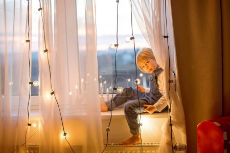 Photo for Toddler child, cute blond boy, sitting on the window, reading book, christmas lights aroundhim - Royalty Free Image