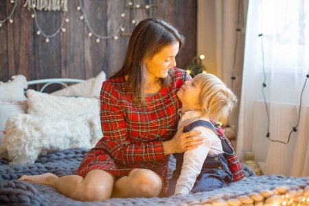 Téléchargez les photos : Enfant tout-petit embrassant la mère au lit, mignon garçon blond et maman embrassant, lumières de Noël autour d'eux - en image libre de droit