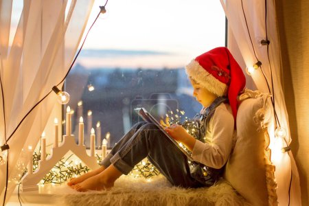 Photo for Toddler child, cute blond boy, sitting on the window, reading book, christmas lights aroundhim - Royalty Free Image