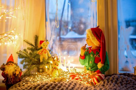 Téléchargez les photos : Mignon tout-petit enfant en pyjama, assis sur une couverture furieuse à côté d'une fenêtre, regardant dehors les feux d'artifice la veille du Nouvel An - en image libre de droit