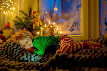 Téléchargez les photos : Mignon tout-petit enfant en pyjama, dormant sur une couverture furieuse à côté d'une fenêtre, nuit bleue à l'extérieur avec feux d'artifice la veille du Nouvel An - en image libre de droit