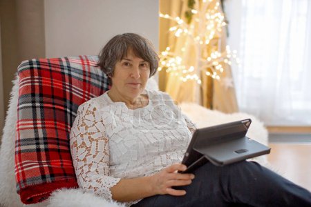 Téléchargez les photos : Elderly woman, sitting on a cozy armchair at home on Christmas, watching tablet, relax - en image libre de droit