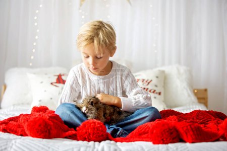 Foto de Hermoso niño rubio, lindo niño, sosteniendo conejo pequeño, abrazándolo en casa - Imagen libre de derechos