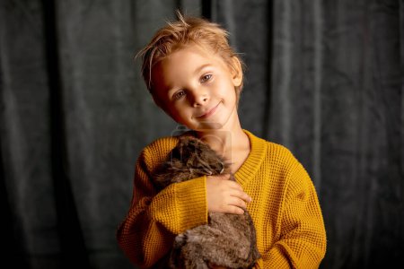 Foto de Hermoso niño rubio, lindo niño, sosteniendo conejo pequeño, abrazándolo en casa - Imagen libre de derechos