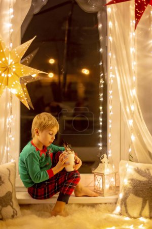 Foto de Toddler child, cute blond boy, sitting on the window, reading book, Christmas lights around him - Imagen libre de derechos