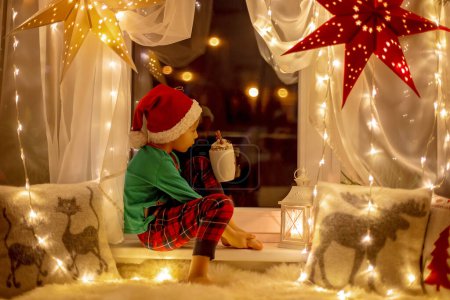Téléchargez les photos : Toddler child, cute blond boy, sitting on the window, reading book, Christmas lights around him - en image libre de droit