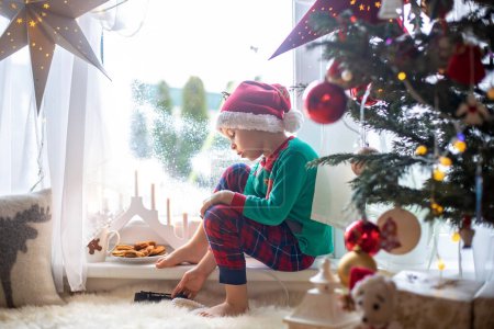 Photo for Toddler child, cute blond boy, sitting on the window in pajama, looking out for Satna Claus,, eating cookies,  Christmas lights around him - Royalty Free Image