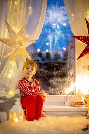 Foto de Toddler child, cute blond boy, sitting on the window in pajama, looking out for Satna Claus,, eating cookies,  Christmas lights around him - Imagen libre de derechos