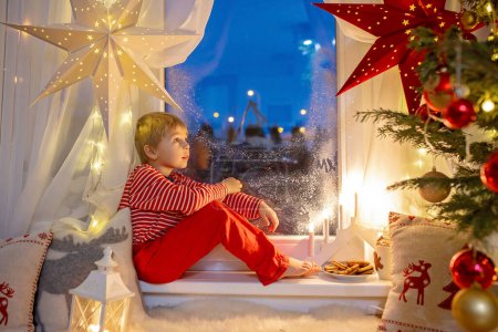 Téléchargez les photos : Toddler child, cute blond boy, sitting on the window in pajama, looking out for Satna Claus,, eating cookies,  Christmas lights around him - en image libre de droit