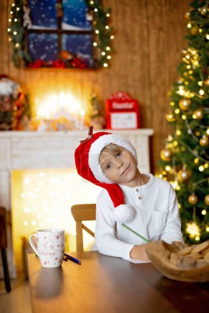 Foto de Hermoso niño rubio, joven escolar, escribiendo una carta a Santa Claus en una casa decorada, bebiendo leche y comiendo galletas. Ligths y juguetes de punto alrededor - Imagen libre de derechos