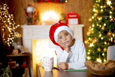 Téléchargez les photos : Belle enfant blonde, jeune écolier, écrivant une lettre au Père Noël dans une maison décorée, buvant du lait et mangeant des biscuits. Ligues et jouets tricotés autour - en image libre de droit