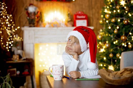 Foto de Hermoso niño rubio, joven escolar, escribiendo una carta a Santa Claus en una casa decorada, bebiendo leche y comiendo galletas. Ligths y juguetes de punto alrededor - Imagen libre de derechos