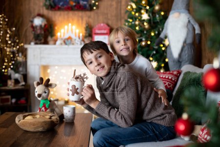 Téléchargez les photos : Beau blond enfant et son vieux frère, jeunes écoliers, jouant dans une maison décorée avec des jouets tricotés à Noël, mangeant des biscuits et buvant du lait - en image libre de droit
