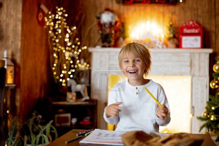 Photo for Beautiful blond child, young school boy, writing letter to Santa Claus in a decorated home, drinking milk and eating cookies. Ligths and knitted toys around - Royalty Free Image