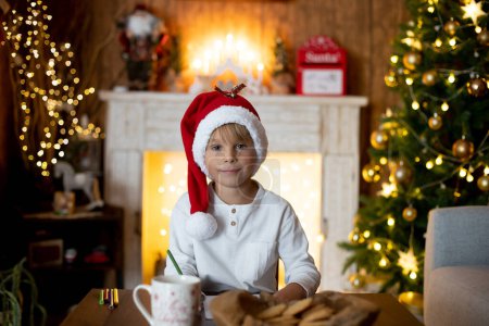 Photo for Beautiful blond child, young school boy, writing letter to Santa Claus in a decorated home, drinking milk and eating cookies. Ligths and knitted toys around - Royalty Free Image