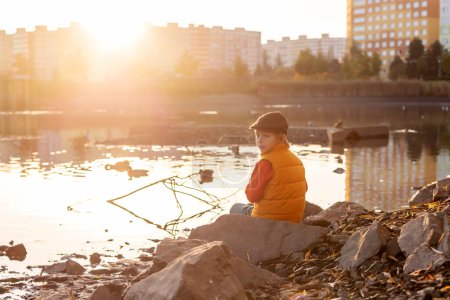 Foto de Hermoso niño rubio preescolar, niño, sentado en el parque en la salida del sol de la mañana, disfrutando del clima de otoño, beber té y disfrutar del sol - Imagen libre de derechos